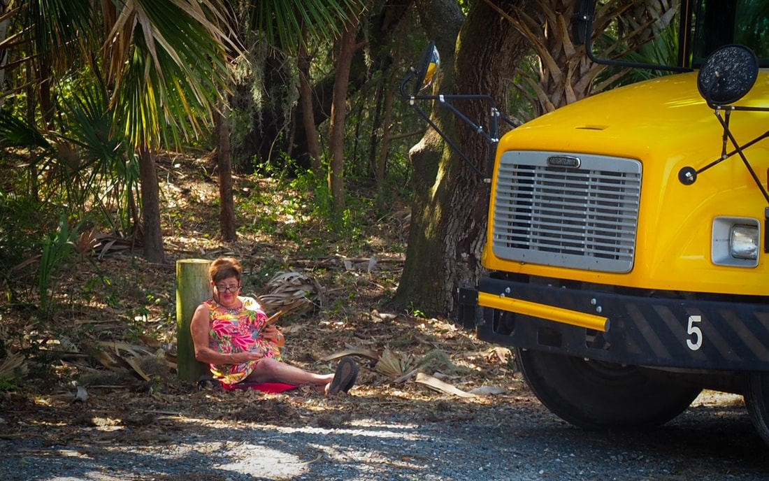 Picture of bus driver sitting on the ground by her bus waiting and reading while the students work on their workbooks